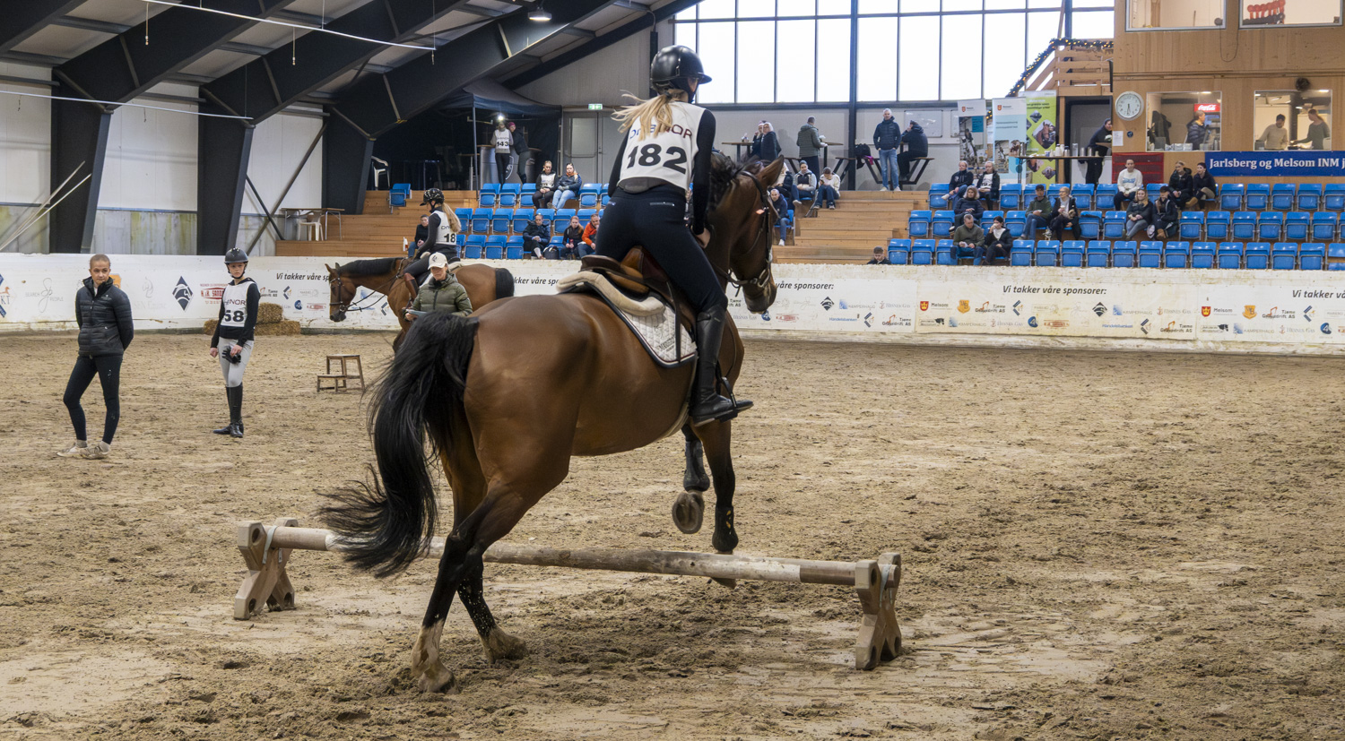 Rytter på hest, hopper over hinder inne i ridehall. Tilskuere.
