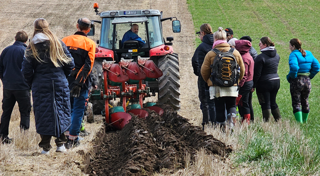 Traktor pløyer på et jorde mens studenter ser på