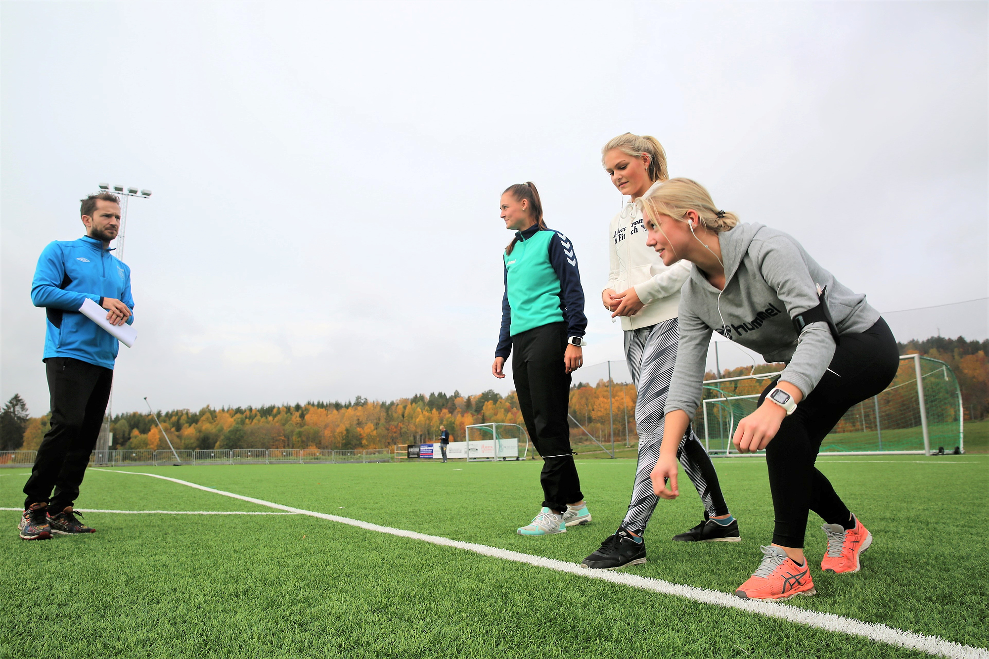 Tre jenter står klare til å løpe på en gressbane. En lærer skal ta tiden.