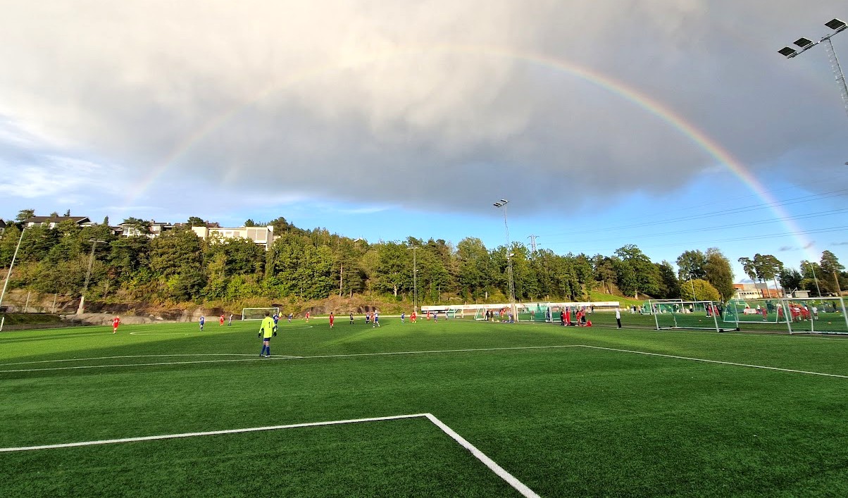 Fotballbane med spillere ute på banen. Regnskyer og regnbue i bakgrunnen.