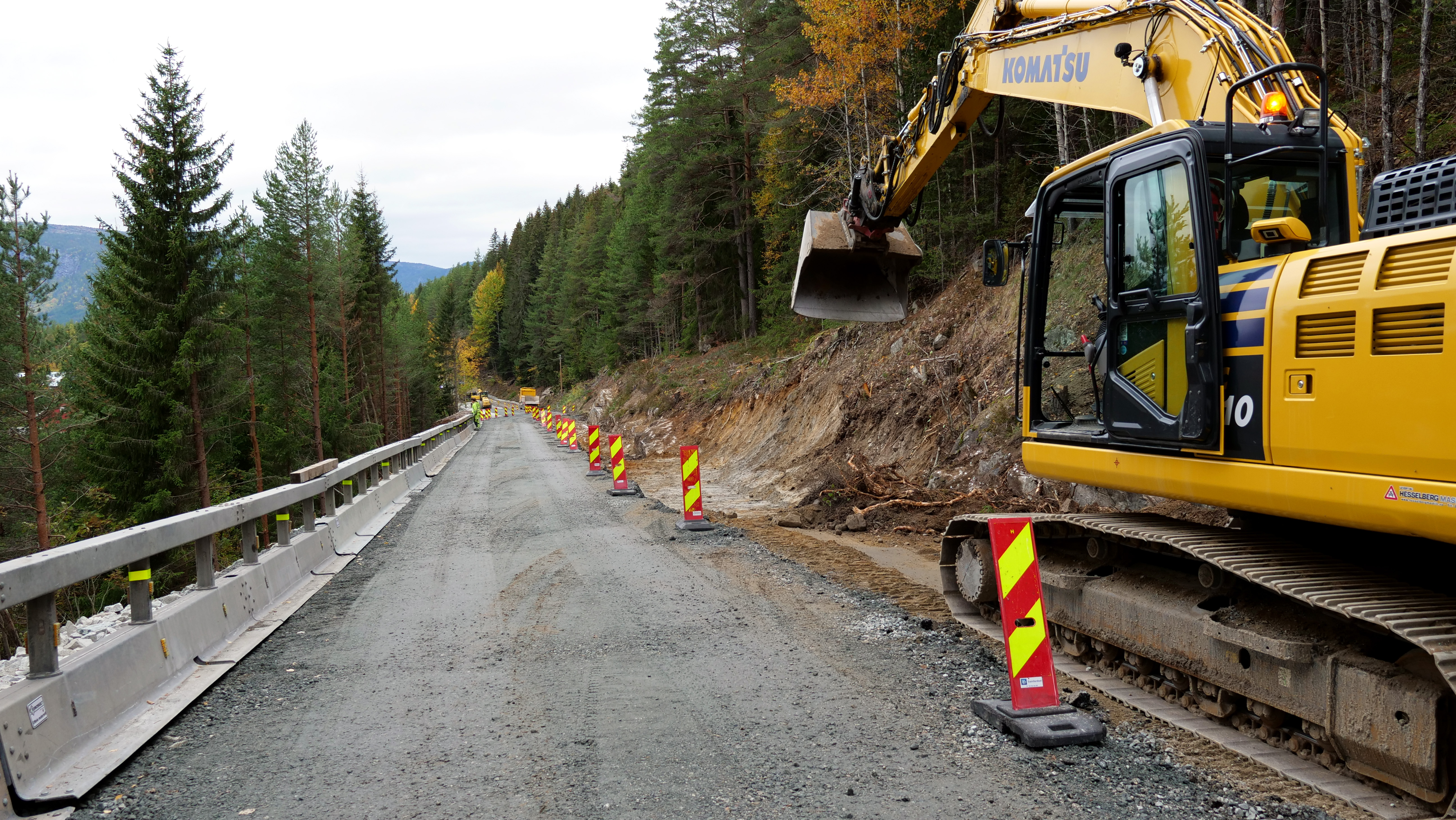 Gruslagt vei går innover i bilde. Gul gravemaskin i arbeid på høyre side.