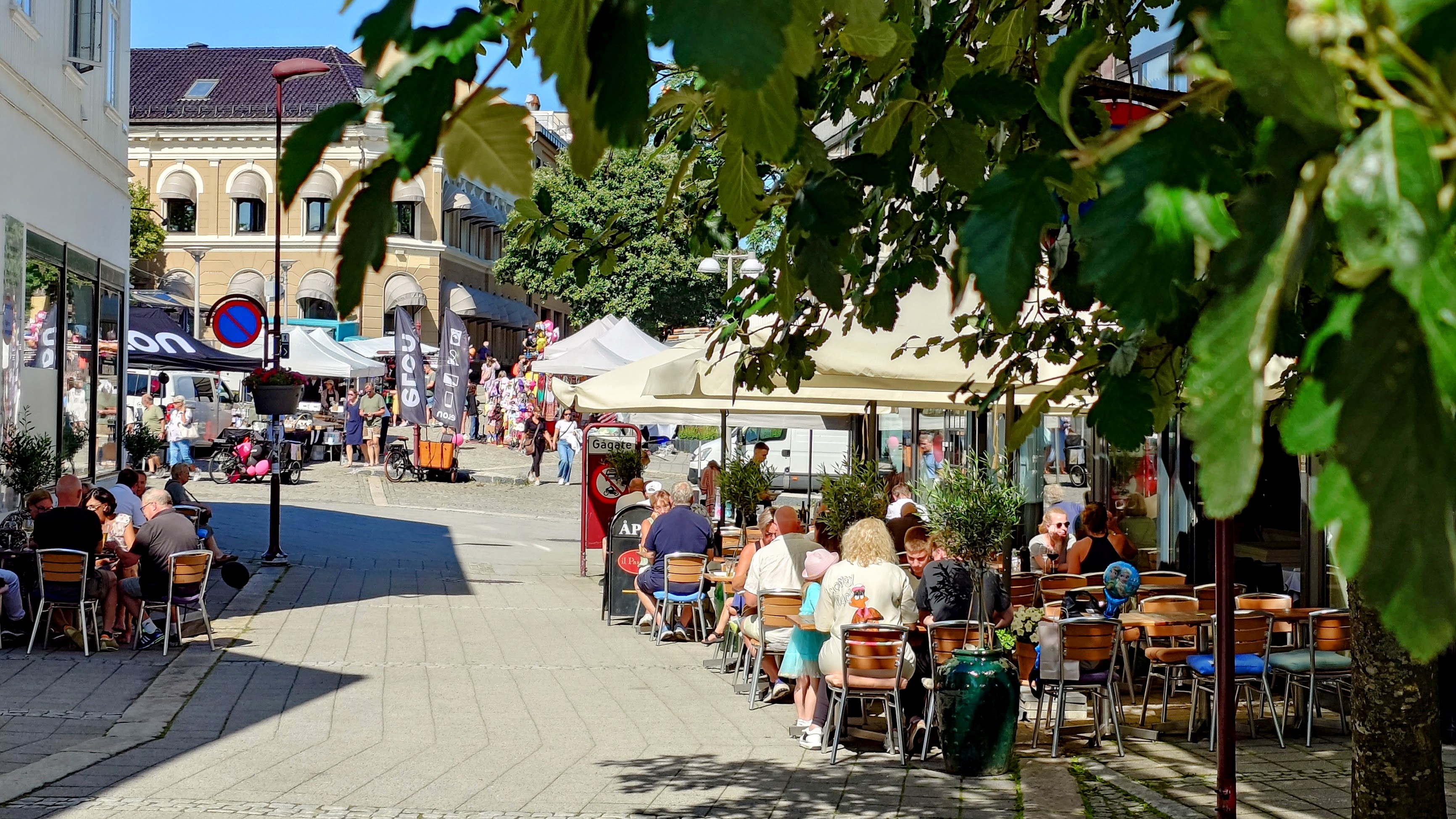 Bildet viser brede gater, grønne trær og folk som spiser på uteserveringene i Larvik.
