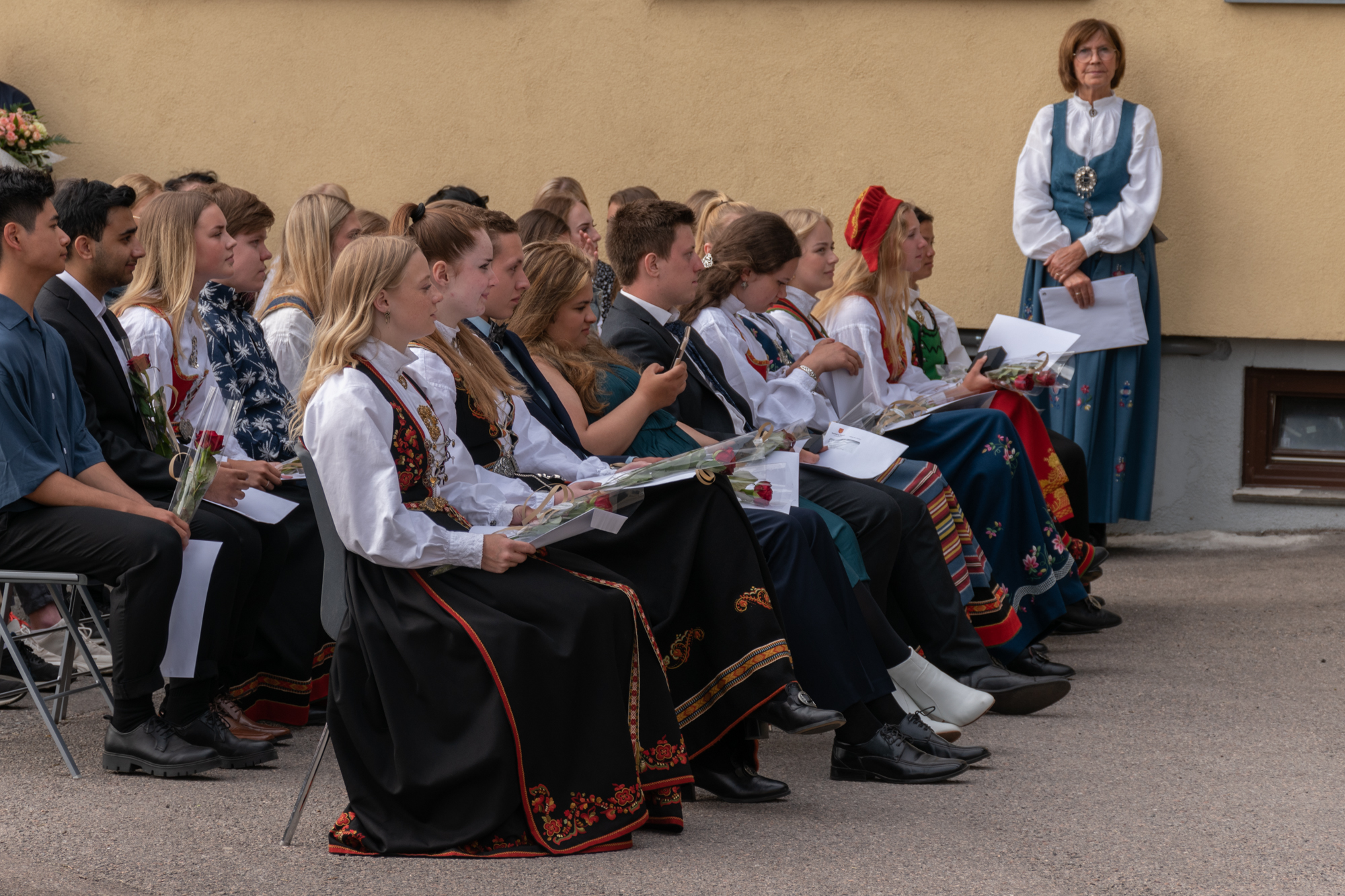 Elever i bunad sitter på stoler ute