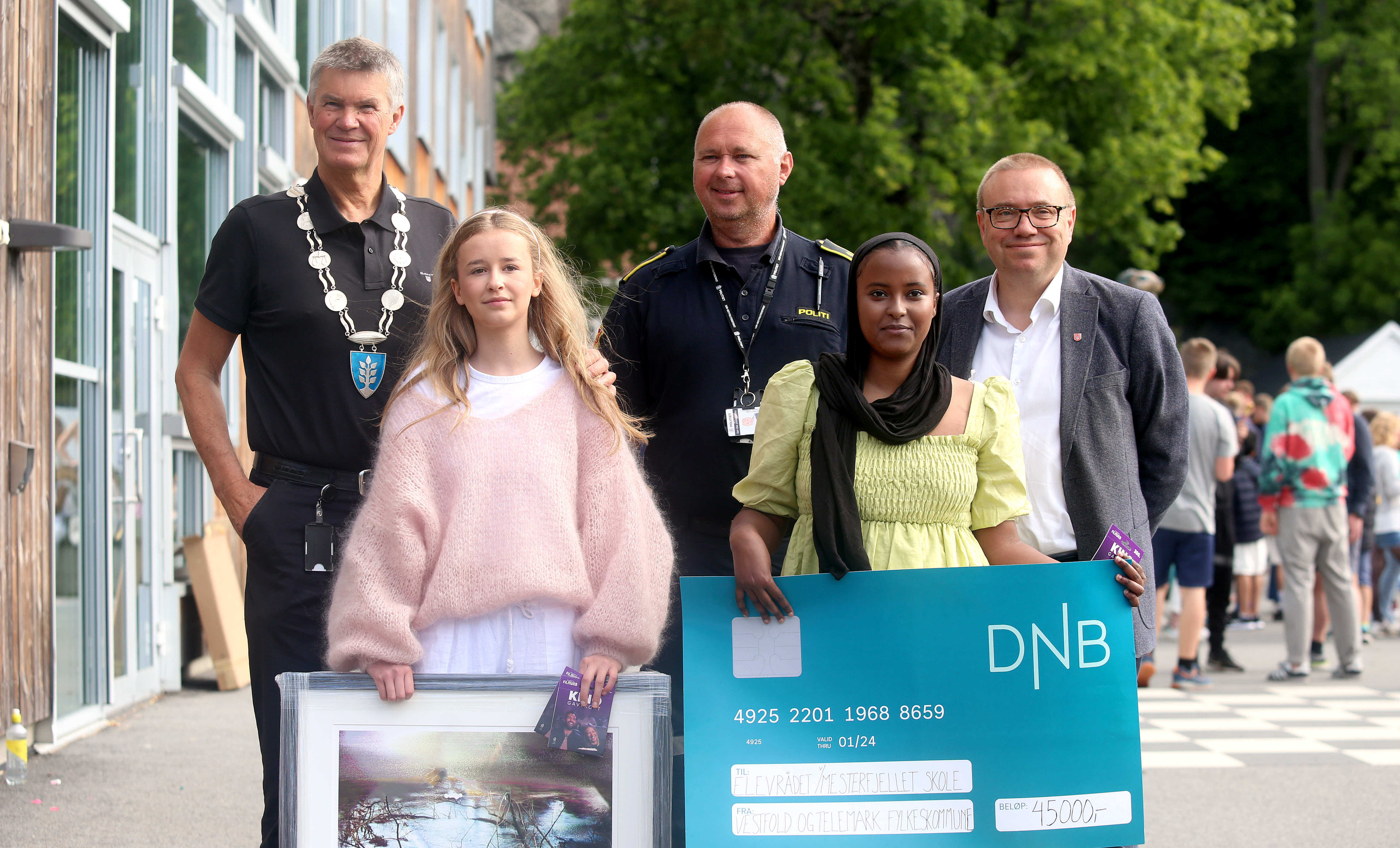 To jenter i forgrunnen av bildet holder et maleri og en sjekk. Tre menn står bak. Alle ser i kamera.