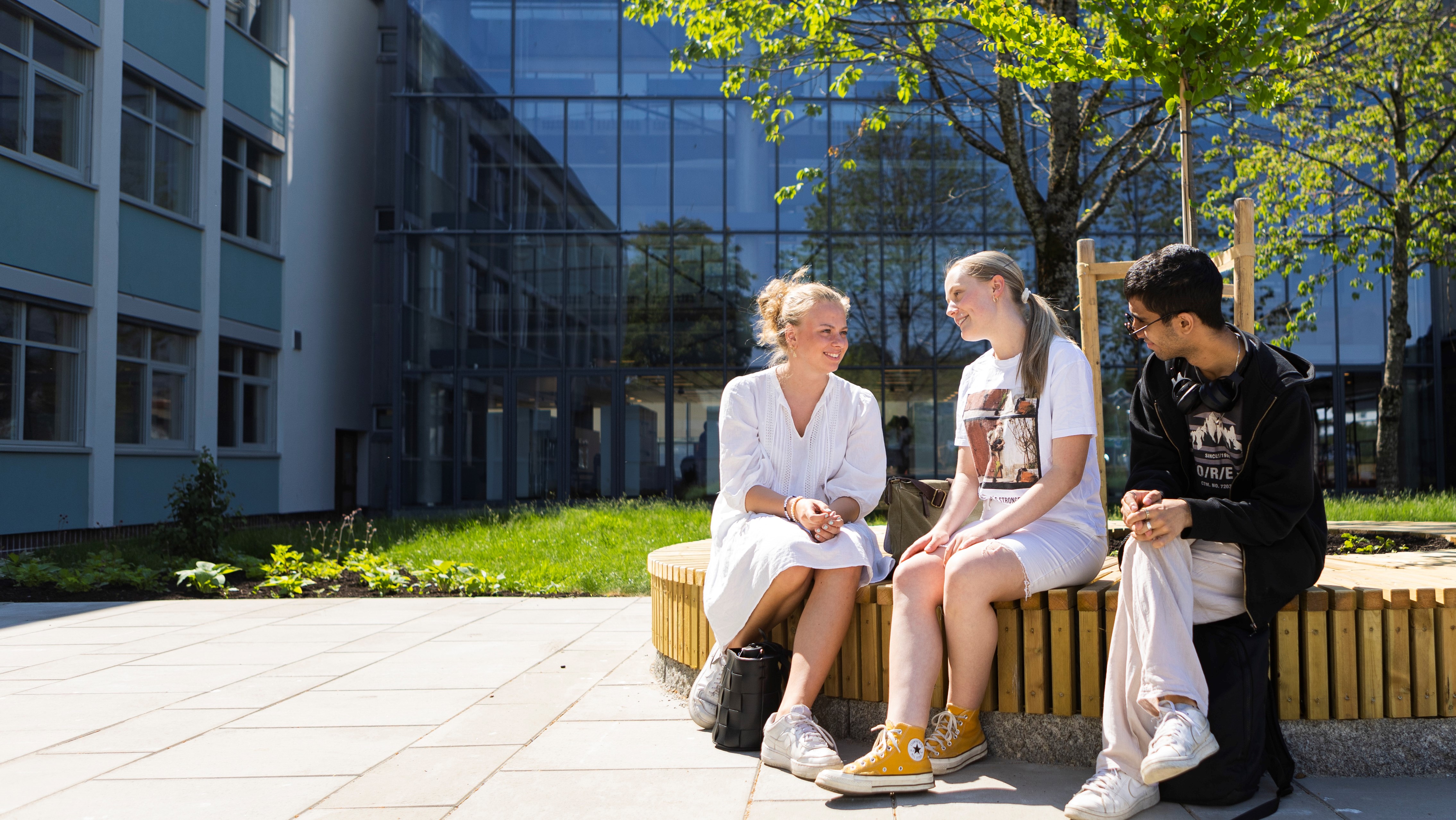To jenter og en gutt sitter på en benk foran skolen.
