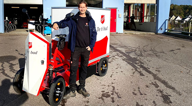 På bildet står Jonas Bogen Berg sammen med den elektriske varesykkelen til Frelsesarmeen. Den har fire hjul og stor firkantet og tett lastekasse bakpå. Den er i fargene rød og hvit.