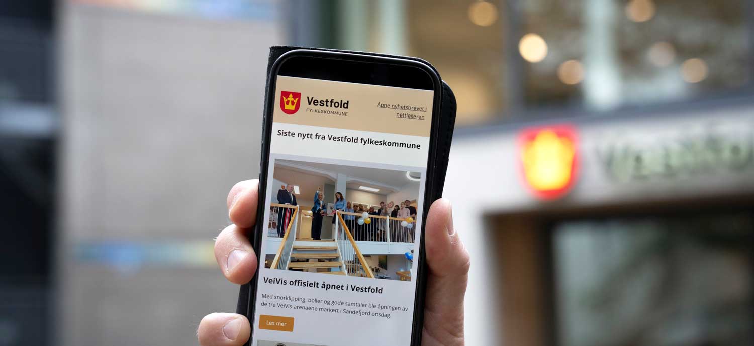 En hånd holder en mobiltelefon som viser et nyhetsbrev fra Vestfold fylkeskommune