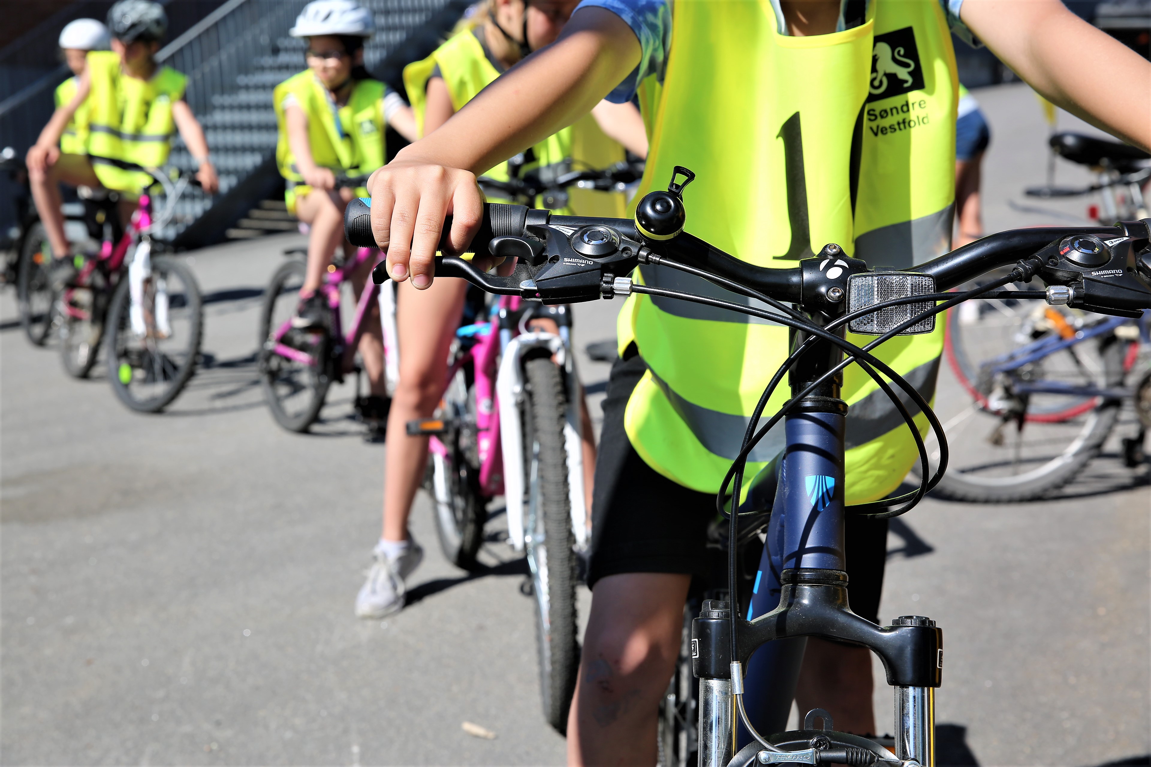 Syklist som vises fra knærne og opp til skuldrene fyller høyre bildeflate. I bakgrunnen skimtes andre syklister i gul vest.